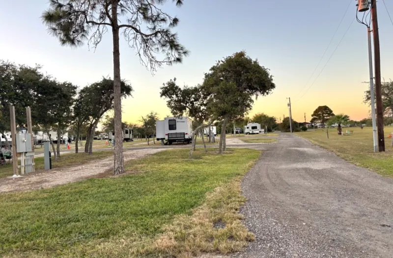 A gravel drive through Bay Breeze RV Park at sunset, with travel trailers parked at their sites and hookup pedestals along the grass