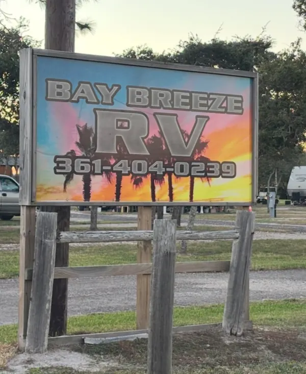 The Bay Breeze RV sign at the park entrance, a painted sunset behind palm trees with the phone number (361) 404-0239